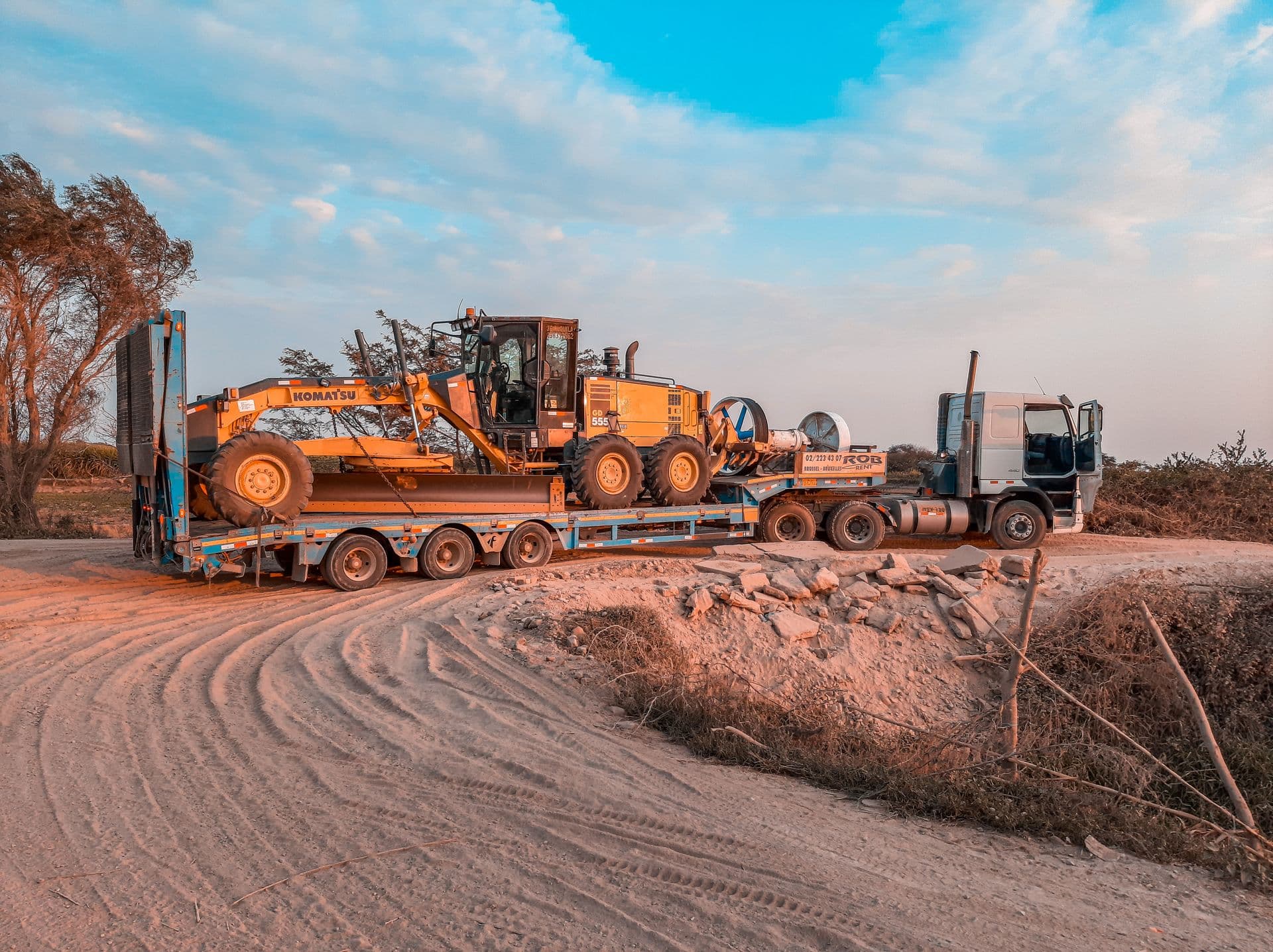 Prime mover hauling a grader on a low-loader trailer along a dusty road